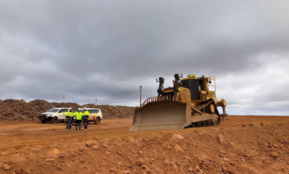 a bull dozer stands in front of workers talking on a red dirt road.